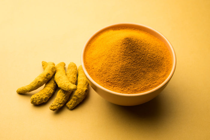 Bowl of turmeric powder with fresh turmeric roots on yellow background, representing the golden spice used in daily wellness rituals