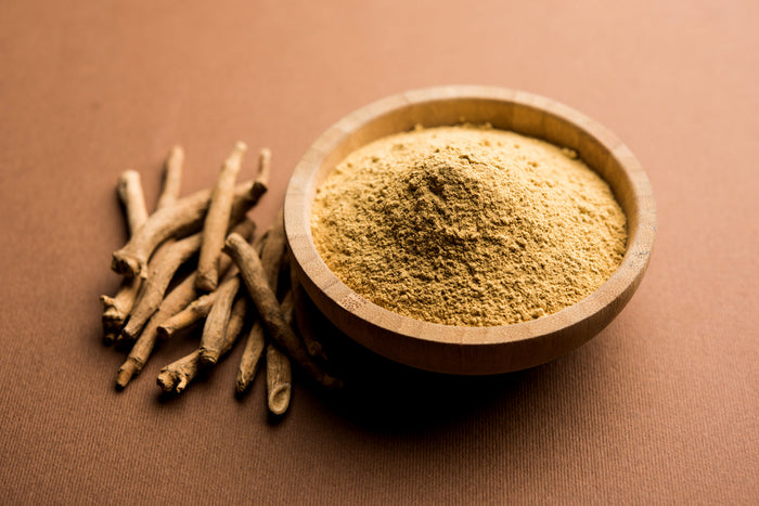 Wooden bowl filled with ashwagandha powder beside dried roots on brown surface, showing traditional herbal preparation