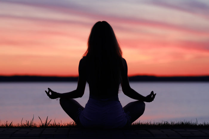 Silhouette of woman meditating at sunset near water, reflecting calmness, stress relief, and mind-body balance