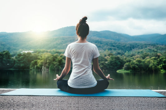 Woman practicing meditation outdoors by a lake with mountains in the background, promoting Ayurveda-inspired warmth, balance, and inner energy during November.