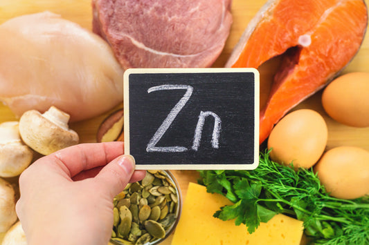 Hand holding a zinc sign in front of zinc-rich foods including salmon, eggs, chicken, seeds, mushrooms, cheese, and herbs
