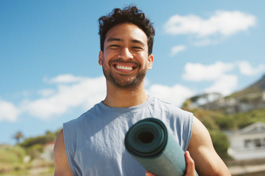 Smiling man holding yoga mat outdoors under a sky, representing happy active wellness, healthy living, and cellular health support each day.